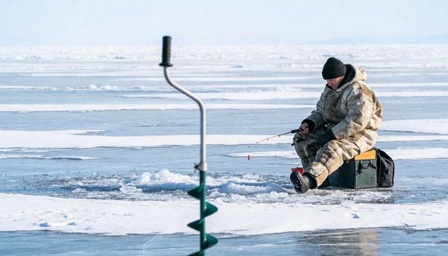 Solitary angler fishing on frozen lake with manual auger and insulated winter gear