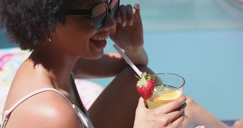 African american woman smiling and holding citrus cocktail with strawberry poolside