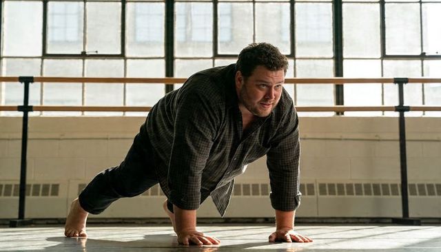 Man holding plank in sunlit rehearsal studio with wooden barre and large windows