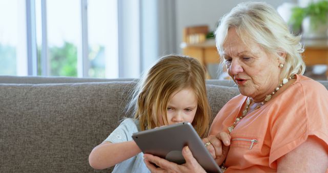 Grandmother sitting with young granddaughter, using tablet together on living room couch. Ideal for showcasing technology use across generations, family bonding, learning, and home education themes.