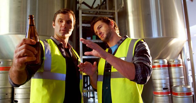 Two male brewery workers in safety vests examining a beer bottle. One is holding the bottle and looking at it closely, while the other is gesturing, possibly explaining or discussing. Large metal brewing tanks and kegs are visible in the background suggesting an industrial environment. This can be used for illustrating concepts related to brewery operations, quality control in manufacturing, teamwork in industrial settings, or training materials in the brewing industry.