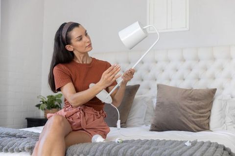 Woman installing white bedside lamp while sitting on bed in cozy neutral bedroom