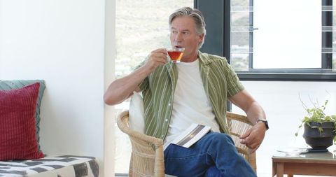 Senior man sipping tea in contemporary bright living room while reading paperback