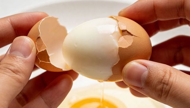 Hands peeling hardboiled brown egg with cracked shell and yolk dripping into bowl closeup