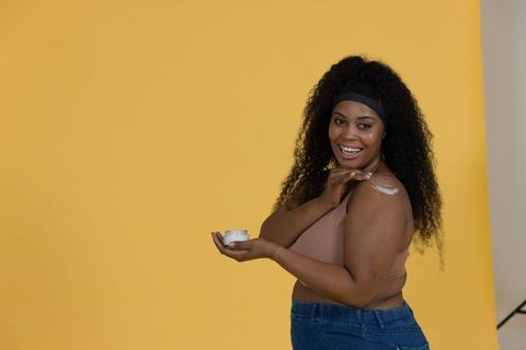Smiling african american woman applying shoulder moisturizer on yellow backdrop