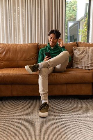 Young asian man relaxing on leather sofa listening with earbuds and using smartphone