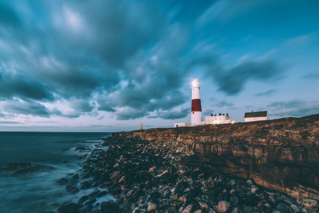 Lighthouse standing tall on a rocky coast with sunset and dramatic clouds covering the sky. Ideal for use in travel blogs, coastal scenery promotions, maritime navigation articles, or inspirational posters.