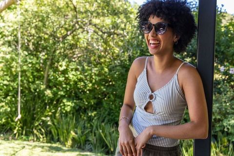 Laughing african american woman leaning on patio post wearing sunglasses and striped top
