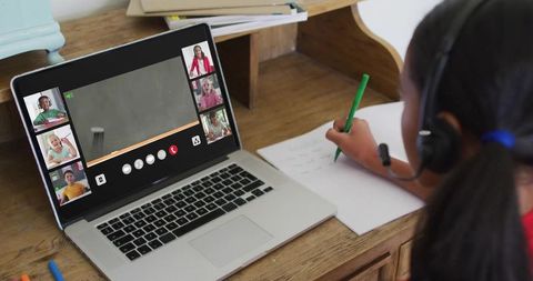 Young student learning remotely at home desk taking notes with laptop headset green pen
