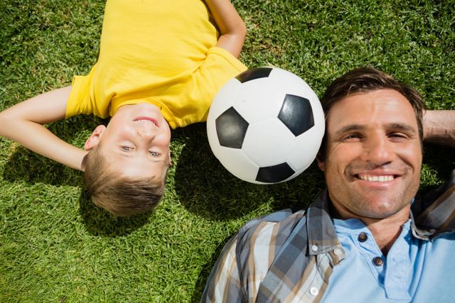Father and son lying on grass in park on sunny day with soccer ball between them. Perfect for themes of family bonding, outdoor activities, leisure time, and parent-child relationships. Ideal for use in advertisements, blogs, and articles about family life, parenting, and summer activities.