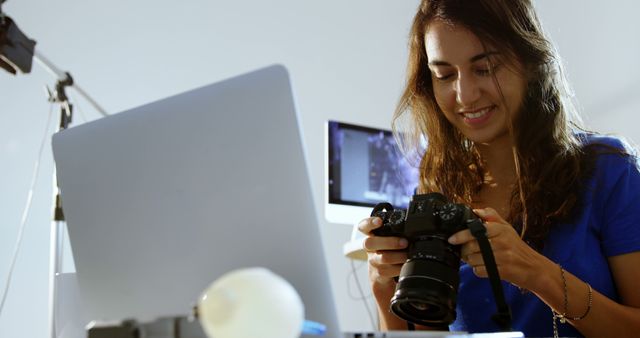 Young woman in a home studio editing photos on laptop, smiling while holding camera. Ideal for illustrations related to freelance work, photography, home office setups, and creative workspaces.