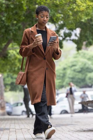 African american woman walking in city wearing brown coat holding coffee and smartphone