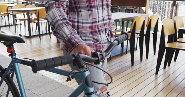 Person dressed casually in a plaid shirt and jeans is holding the handle of a bicycle indoors in a cafe with no other people around. The setting is modern with wooden flooring and yellow chairs. Perfect for promoting urban lifestyle, modern fashion, or active commuting.