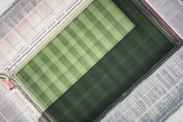 Overhead perspective of a meticulously maintained soccer stadium featuring a crisply mowed field with distinct green stripes. Appropriate for use in sports-related materials, advertisements, promotional campaigns for events, and showcasing sports facilities. Suited for articles on stadium design, sportsground maintenance, or football culture.