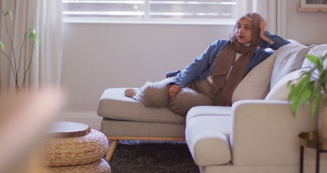 Image of smiling biracial woman in hijab relaxing on sofa at home. Relaxation, happiness, inclusivity and domestic life.