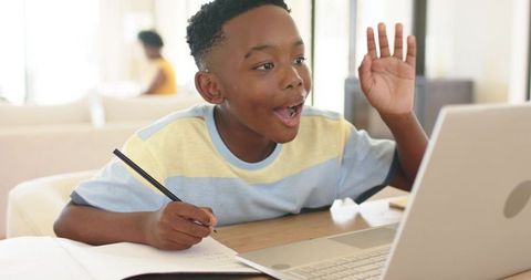 African American boy studying and waving during online class in living room with laptop
