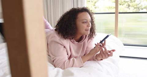 African american woman reclining on white bed using smartphone in bright bedroom