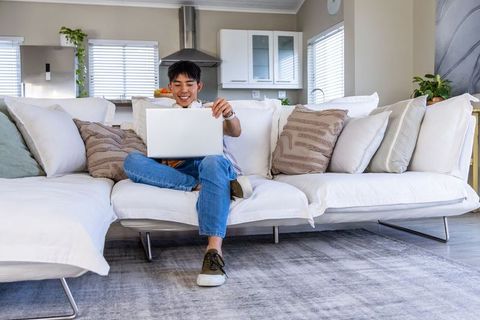 Teenage asian male using laptop on white sectional couch in modern bright living room