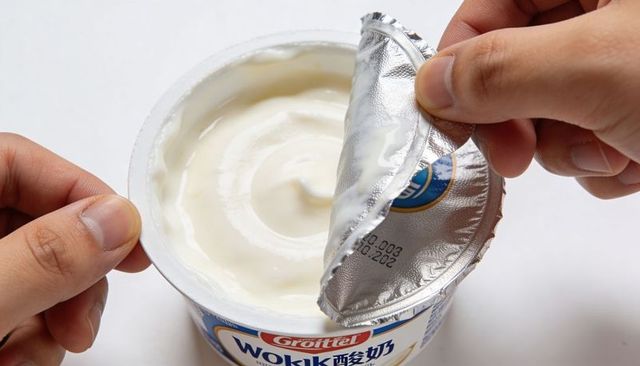 Hands peeling foil lid from yogurt cup revealing creamy white yogurt and label