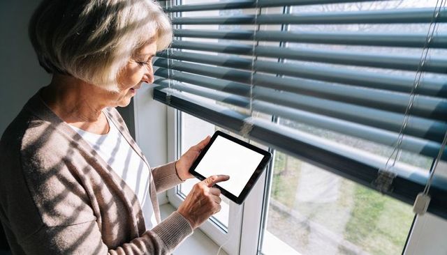 Senior woman using tablet by sunlit window with blinds and charger, tapping touchscreen