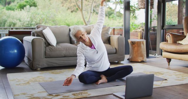 Senior woman practicing yoga in living room with an online class on laptop. Ideal for content related to healthy aging, remote fitness classes, maintaining flexibility and strength, and technology aiding home workouts.