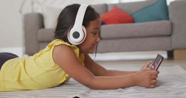 Girl wearing headphones, focusing on a smartphone with a bright screen. She is laying on the floor in a cozy living room environment, suggesting a relaxed and joyful atmosphere. Useful for illustrating children's engagement with technology, enjoyment of music and games. Ideal for educational, recreational, and lifestyle content depicting modern technological usage by children.