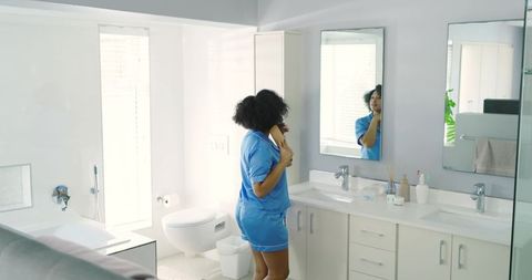 African American woman in blue scrubs brushing hair at modern bathroom vanity
