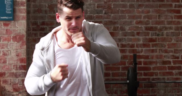 A young Caucasian man is throwing a punch towards the camera, showcasing a boxing stance, with copy space. His intense expression and dynamic pose suggest a moment of action or a training session in a gym with a brick wall background.