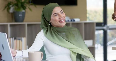 Indian woman in green hijab smiling while working at laptop in bright modern office