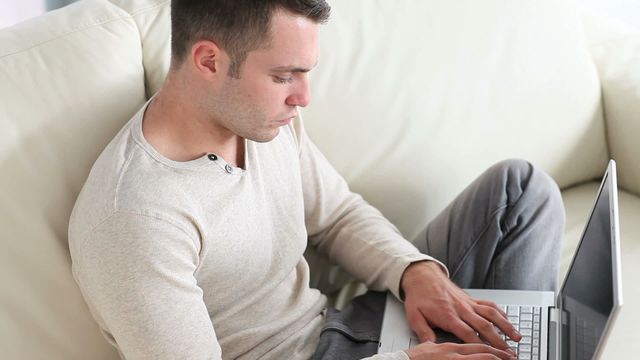 Young man sitting comfortably on a white sofa deeply engrossed in typing on a laptop. Useful for illustrating themes of remote work, home office setups, concentration, and productivity. Perfect for articles, blogs, and ads related to freelancing, at-home work, or comfortable living spaces.