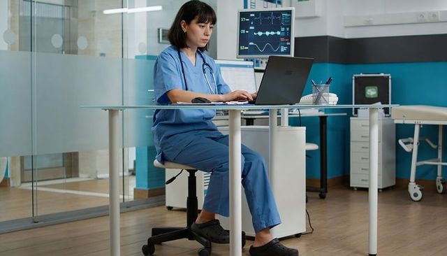 Nurse typing on laptop with ecg monitor in clinic workspace, telemetric monitoring and documentation