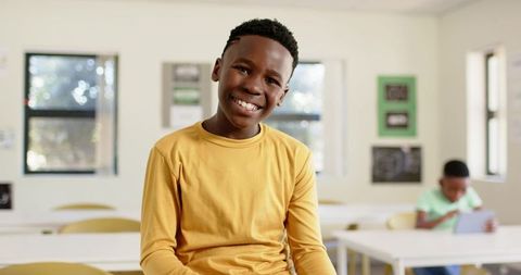 Smiling student in mustard shirt posing in bright classroom while classmate using tablet