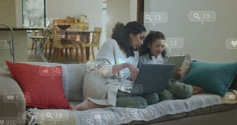Mother and daughter browsing tablet and laptop on sofa with floating social media icons