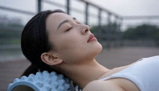 Asian woman relaxing on lakeside boardwalk using blue foam roller for breathwork wellness