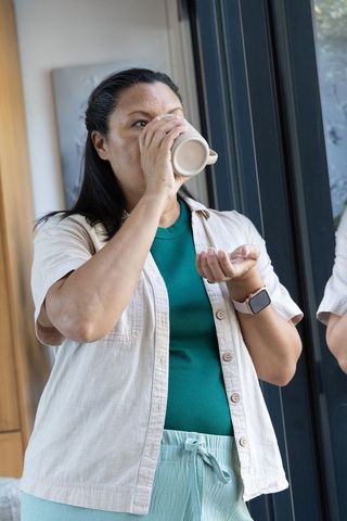 Mature african american woman standing by glass sliding door sipping coffee from ceramic mug