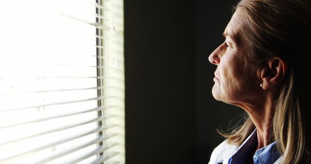 Elderly woman gazing out of window with thoughtful expression. Useful for themes of contemplation, aging, solitude, mental health, and senior lifestyle. Can be used in articles related to senior care, loneliness in old age, reflecting on life, mental wellness, and personal stories.