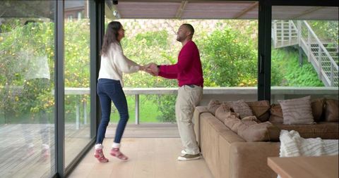 Couple holding hands and dancing in bright modern living room by glass doors