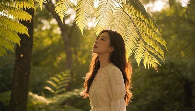 Standing asian woman gazing beneath sunlit fern fronds in backlit forest wearing lace dress