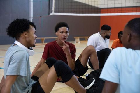 African american volleyball teammates sitting and talking on indoor court wearing knee pads