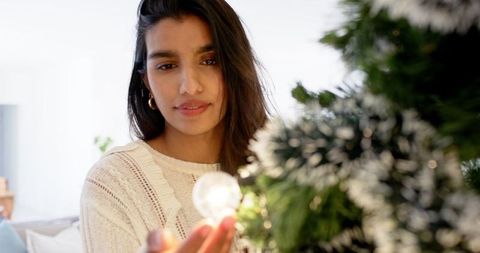 Young woman decorating christmas tree with lights in cozy living room