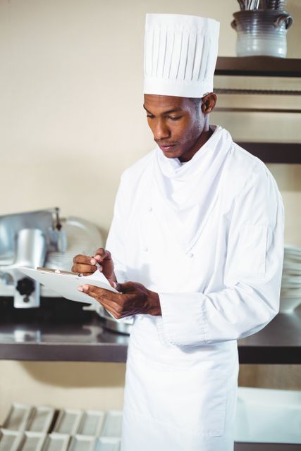Professional chef writing notes on a clipboard in a kitchen. Ideal for use in culinary blogs, restaurant websites, cooking tutorials, and food industry publications. Highlights the importance of organization and attention to detail in professional cooking environments.