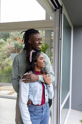 Diverse couple embracing by open sliding door, smiling while looking at garden view