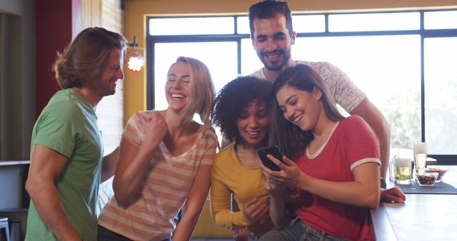 Group of diverse friends are gathered together and enjoying a fun moment while looking at a phone screen. They are standing indoors with happy and relaxed expressions, creating a lively and joyful atmosphere. Perfect for use in content related to friendship, social interactions, happiness, and modern technology.