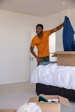 Man packing clothes in bedroom amidst moving boxes in modern apartment