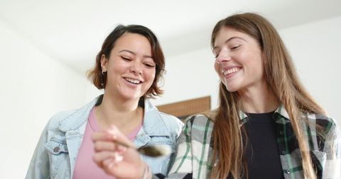 Smiling female friends holding spoon sharing cozy moment in bright minimalist home kitchen