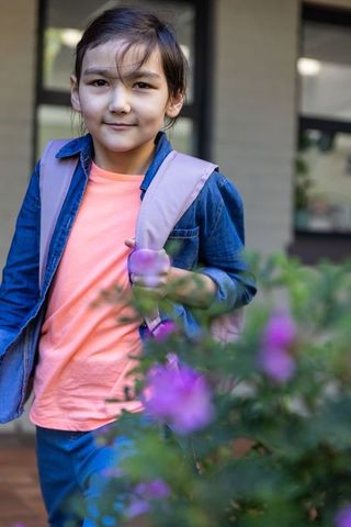 Asian girl standing on porch holding light pink backpack strap wearing denim jacket