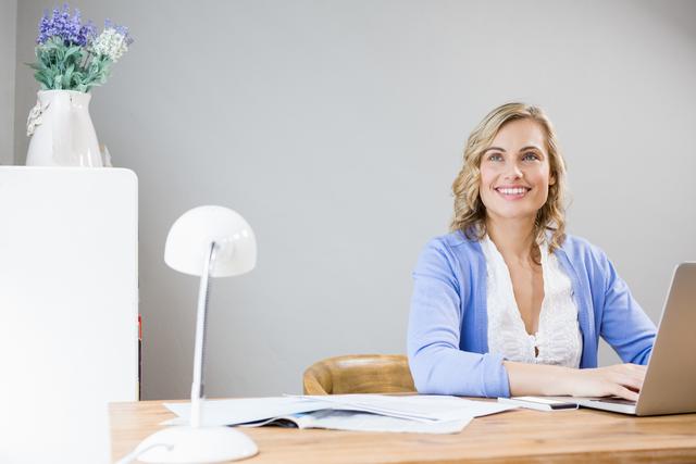 A woman is sitting at her desk, using a laptop and smiling. She appears to be working from home in a relaxed and pleasant environment. This image is perfect for depicting remote work, home office setups, or promoting a work-life balance. It can be used in advertisements, blog posts, or websites focusing on business, technology, or lifestyle themes.