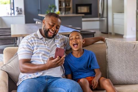 African american father and son laughing and bonding while looking at smartphone on sofa