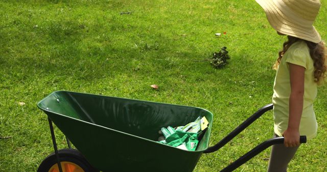 Little girl wearing sun hat pushing wheelbarrow on green lawn. Ideal for promoting children's outdoor activities, gardening, healthy lifestyle, and family time in nature.