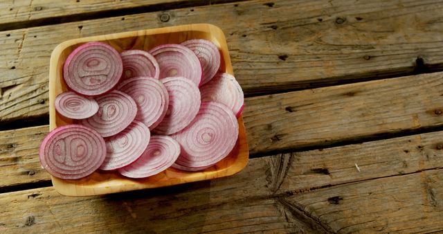 Sliced red onions are neatly arranged on a wooden plate, set against a rustic wooden table background, with copy space. Fresh ingredients like these are essential for adding flavor and color to a variety of culinary dishes.